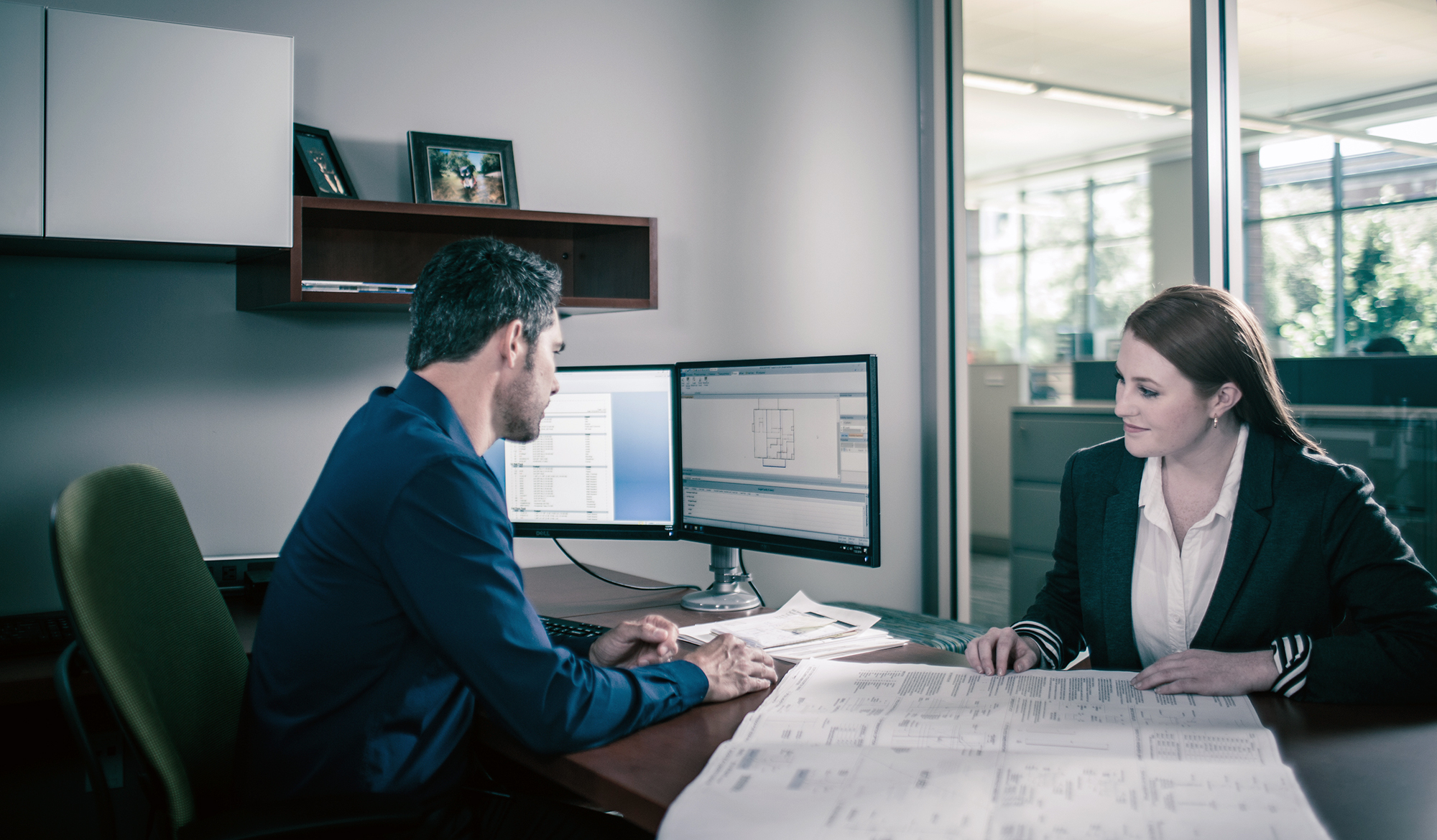 Solutions et services offerts par MiTek - Un homme et une femme sont assis l'un en face de l'autre à un bureau et sur un ordinateur et quelques dessins imprimés.